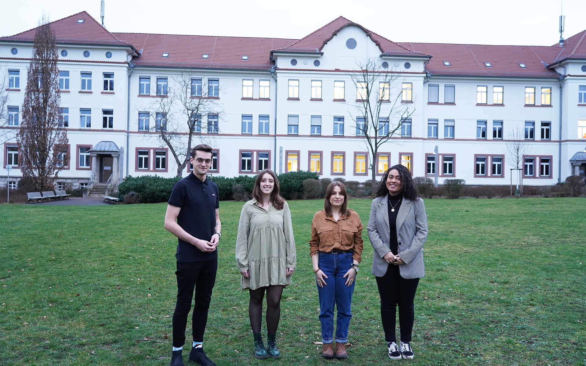 Drei Frauen und ein Mann stehen auf einer Wiese vor einem weißen Gebäude.