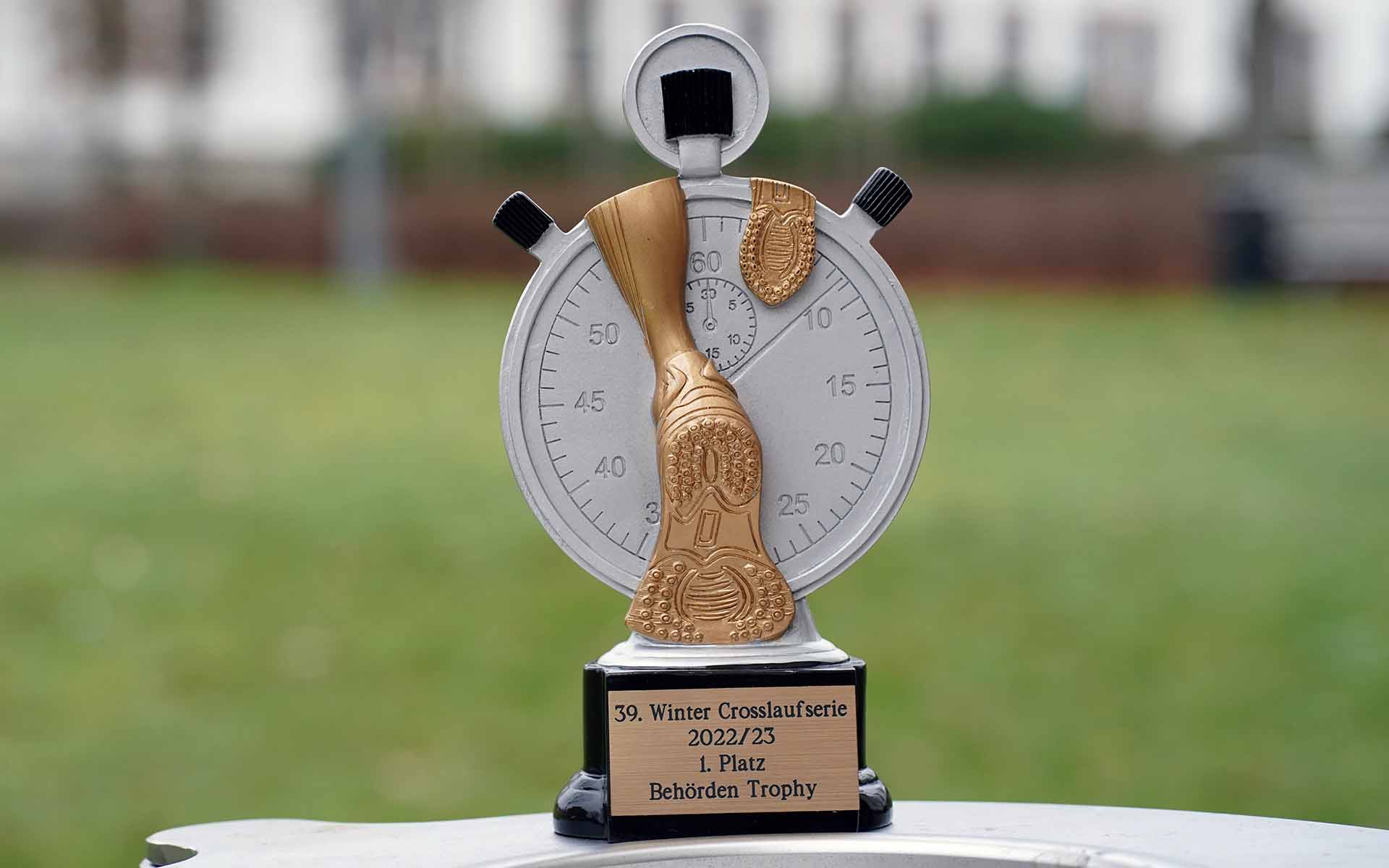 Pokal in Form einer weißen Stoppuhr mit goldenen Laufschuhen auf einem schwarzen Sockel mit graviertem Messingschild vor der grünen Campuswiese