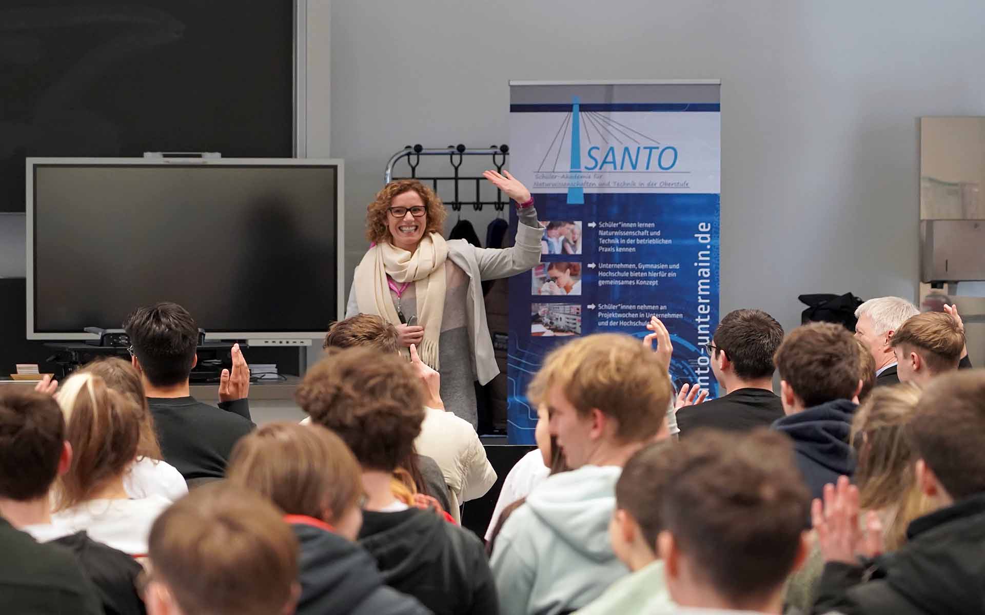 Professorin McNamara steht lächelnd vor den sitzenden Schülerinnen und Schülern und hebt die Hand. Einige Schüler melden sich. Im Hintergrund ist ein blau-weißes SANTO-Roll-up zu sehen.