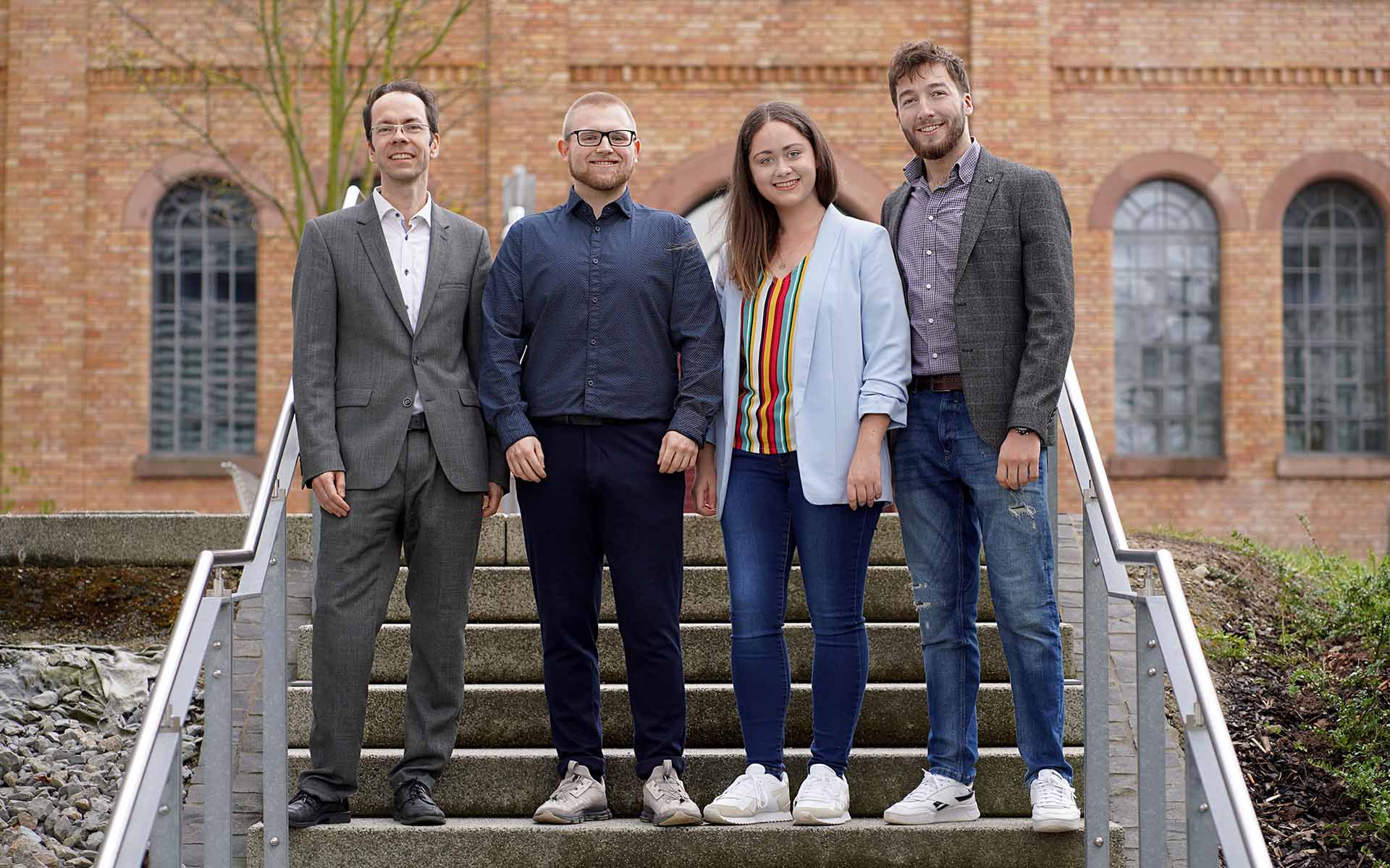 Professor Warnat, Elias Mende, Aileen Reusing und Ismat Zarbaliyev stehen nebeneinander auf einer Treppe vor einem gelb-braunen Backsteingebäude der Hochschule