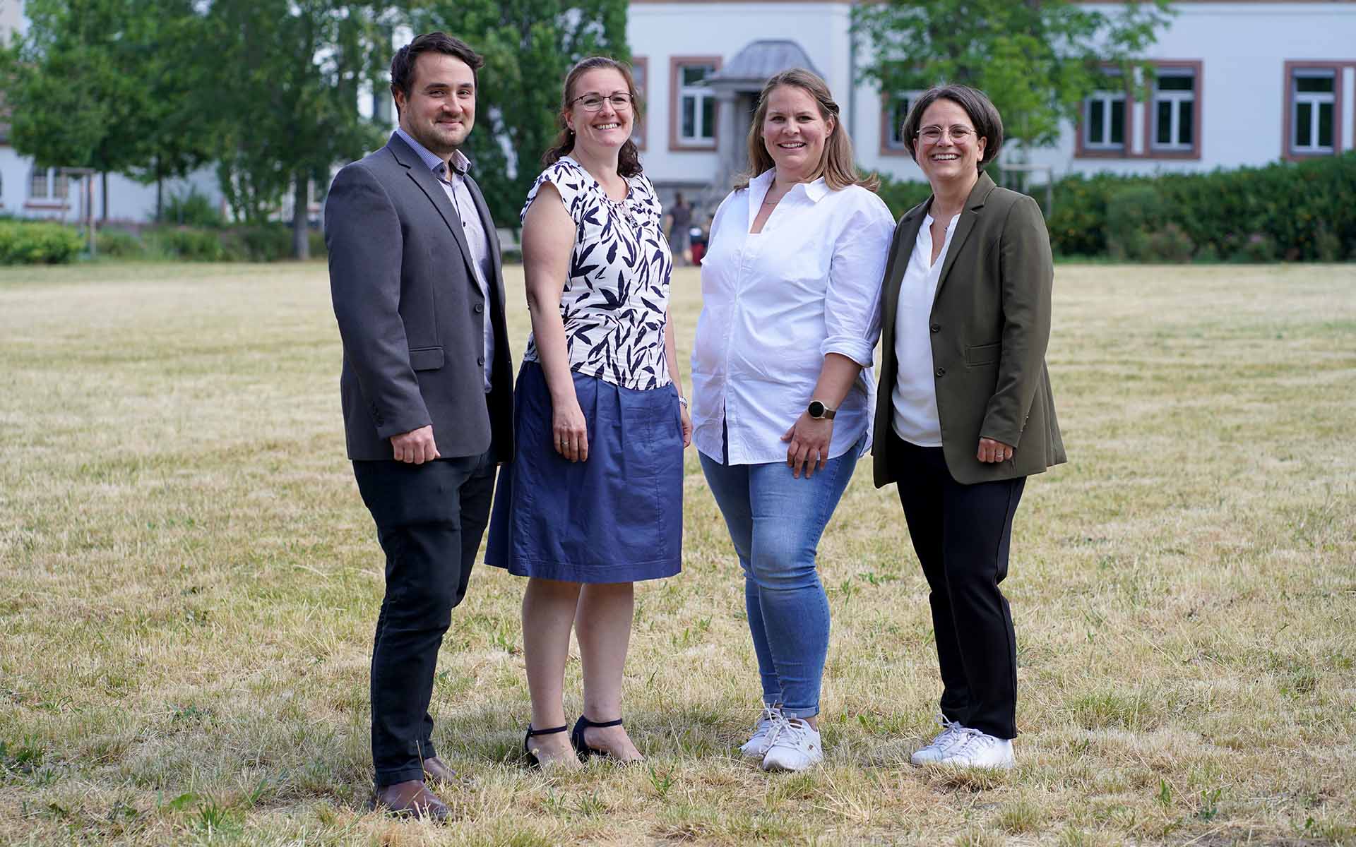 Drei Frauen und ein Mann stehen auf der Campuswiese mit einem weißen Hochschulgebäude und Bäumen im Hintergrund