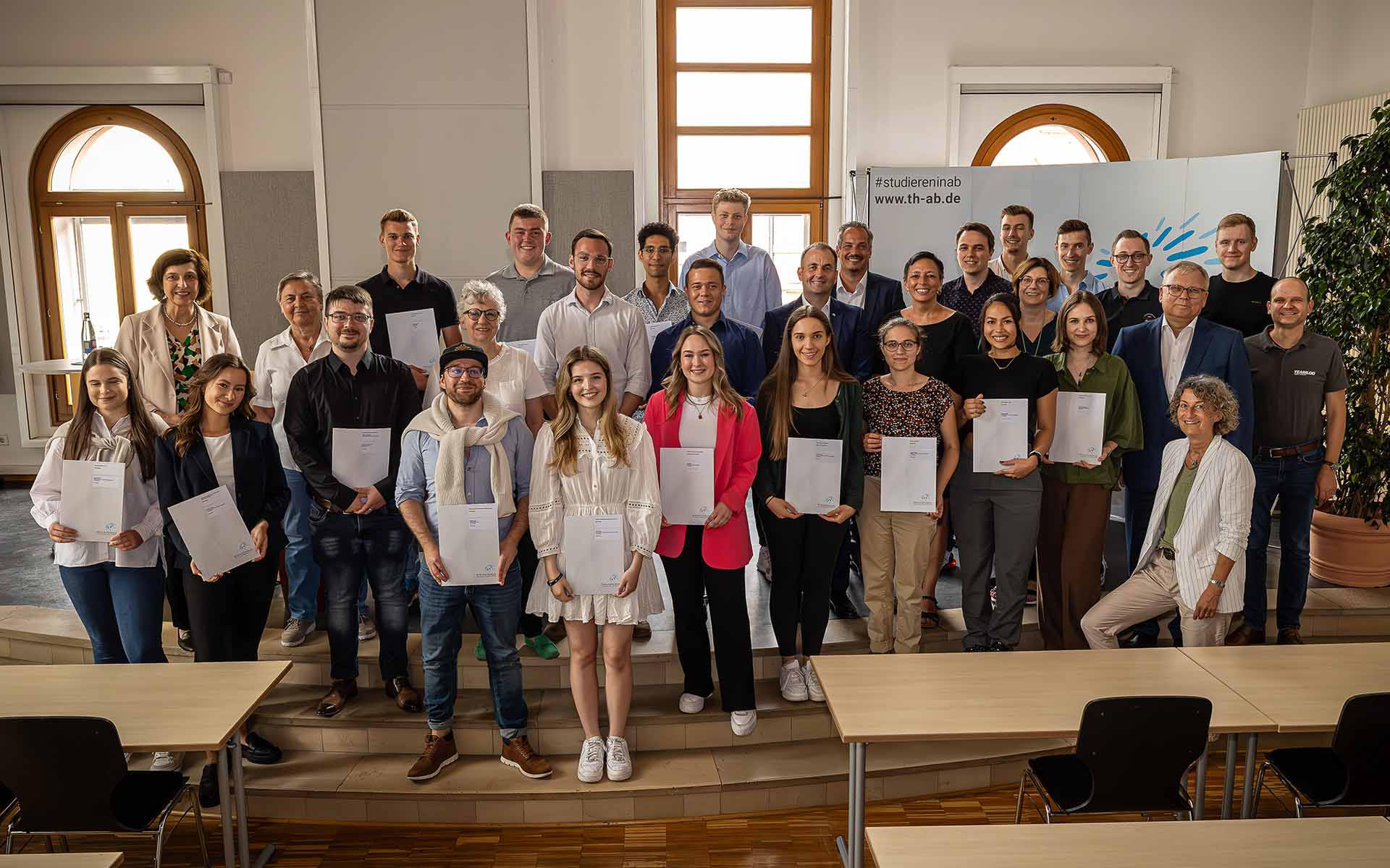 Fördernde, Stipendiatinnen und Stipendiaten stehen in der Aula in mehreren Reihen hintereinander. Einige Personen halten ihre Urkunden in den Hand.