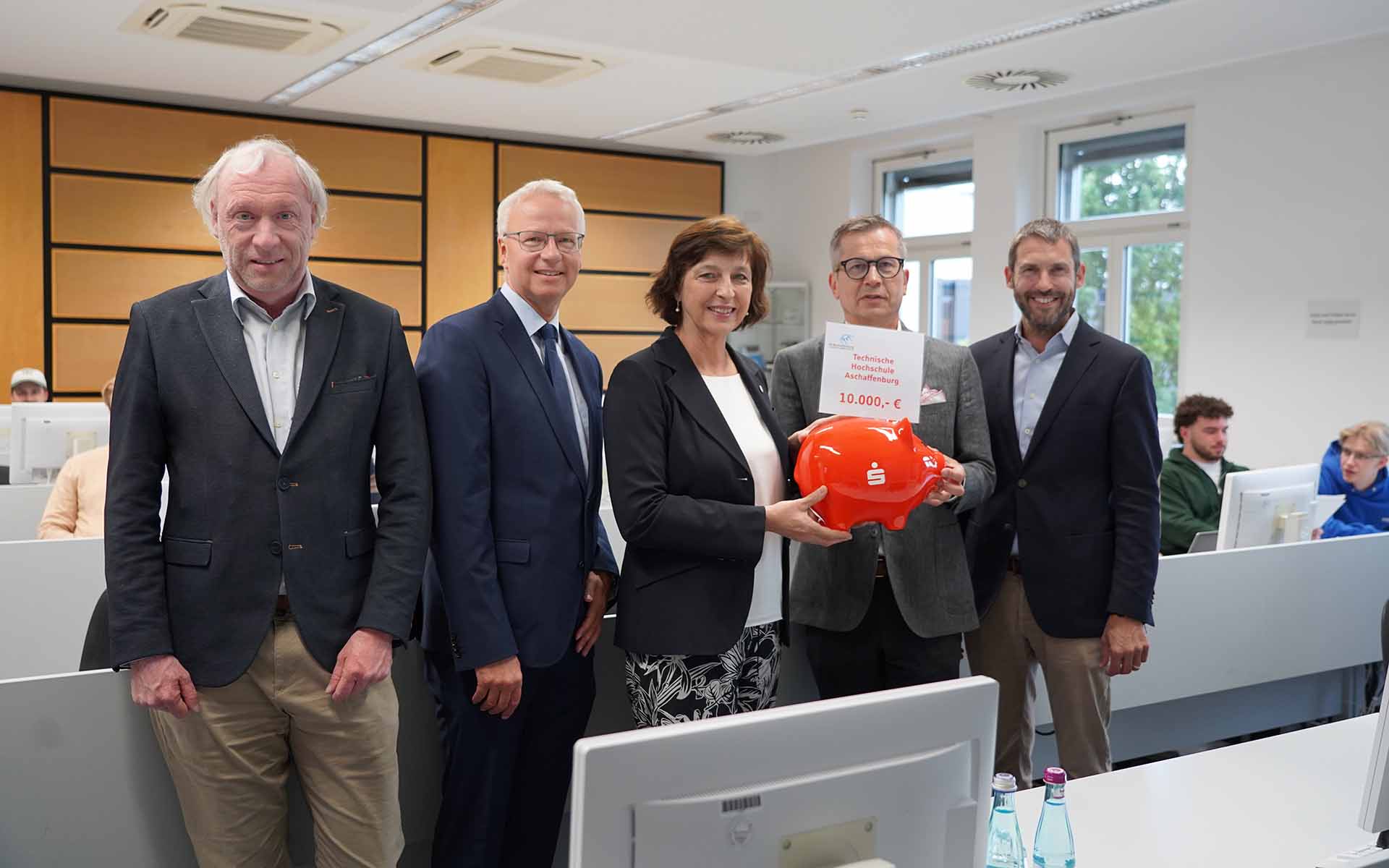 Die Präsidentin der TH AB nimmt das rote Sparschwein mit einem Schild, auf dem die Spendensumme steht, vom Vorstandsvorsitzenden der Sparkasse entgegen. Neben beiden stehen der Landrat, der Laborleiter und der Vizepräsident mit Studierenden an Rechnerarbeitsplätzen im Hintergrund