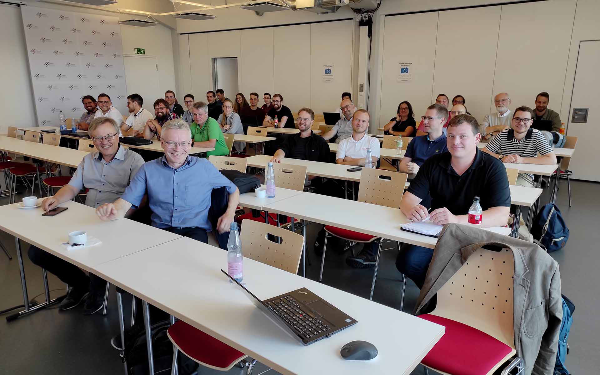 Die Teilnehmenden des Kolloquiums sitzen in einem Hörsaal an Tischen, die in mehreren Reihen hintereinander stehen und blicken lächelnd in die Kamera