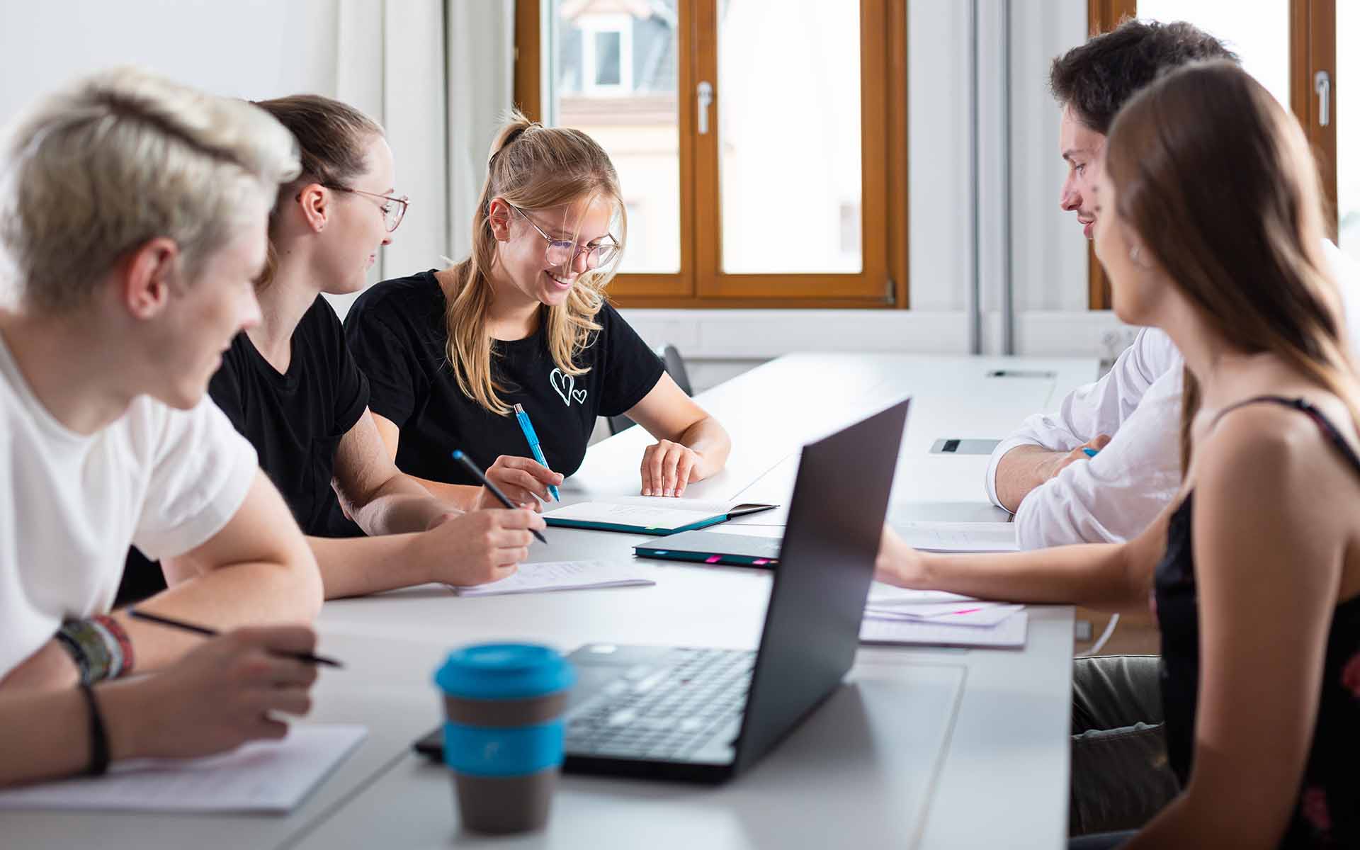 Zwei Studenten und drei Studentinnen sitzen um einen Tisch herum vor einem aufgeklappten Laptop, drei von ihnen haben Stifte in der Hand und Blöcke vor sich liegen