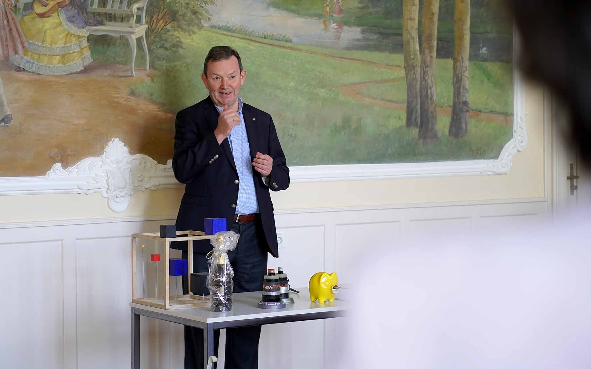 Professor Mußenbrock steht im dunklen Anzug hinter einem Tisch mit verschiedenen symbolischen Gegenständen, anhand derer er wichtig Meilensteine seiner Arbeit an der Hochschule veranschaulicht