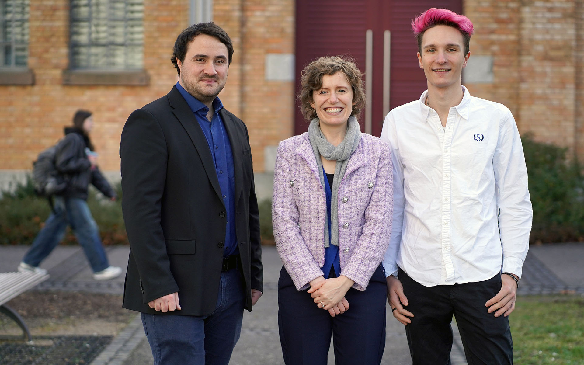 Das Lehrlaborteam der TH Aschaffenburg, bestehend aus einem Mitarbeiter, einer Professorin und einem Studenten, steht vor einem Bachsteingebäude auf dem Campus. Im Hintergrund ist eine vorbeilaufende Studentin zu sehen.