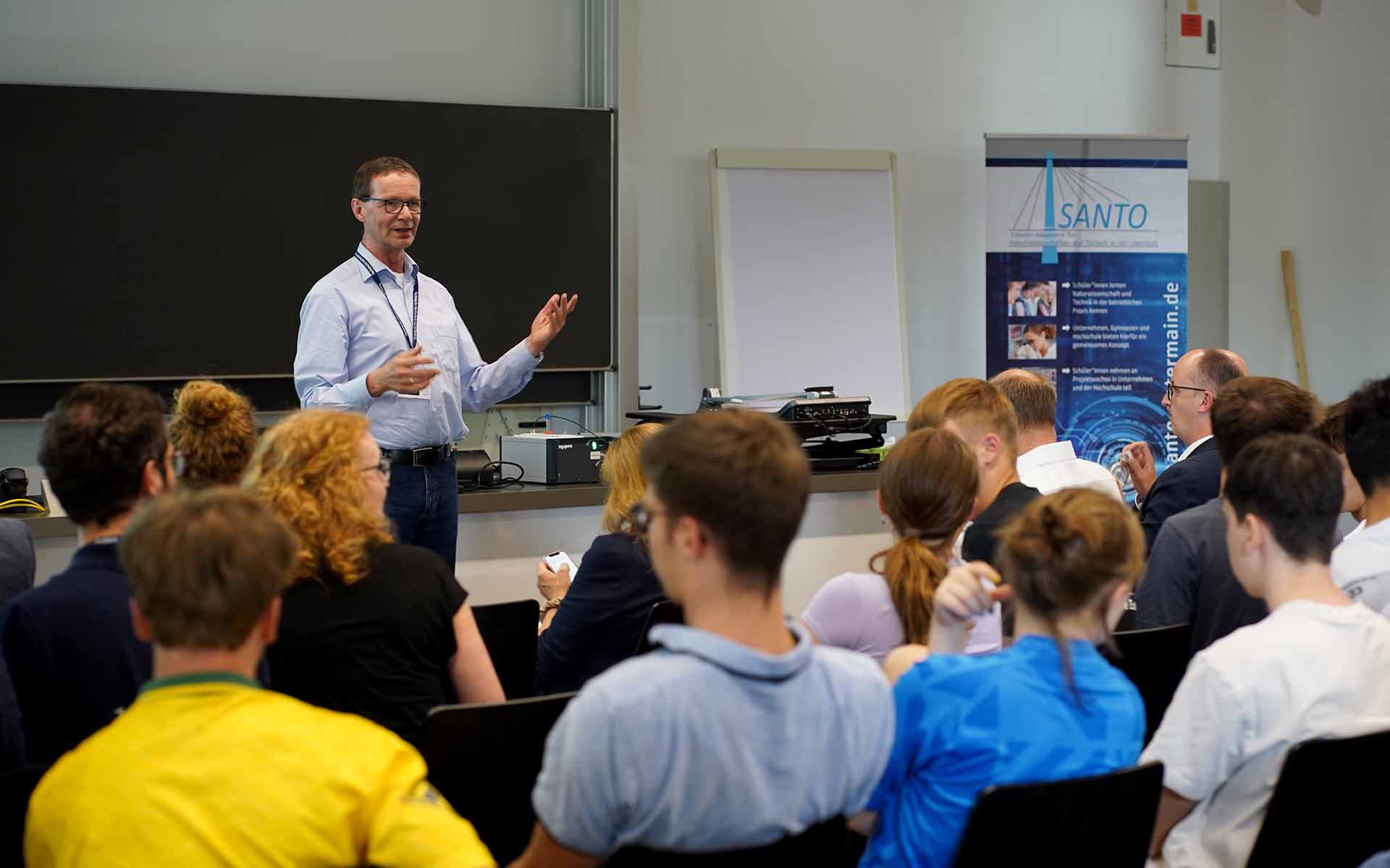 Projektleiter Professor Mewes spricht zum sitzenden Publikum im Hörsaal vor einer Tafel mit einem blau-weißen SANTO-Rollup rechts im Hintergrund