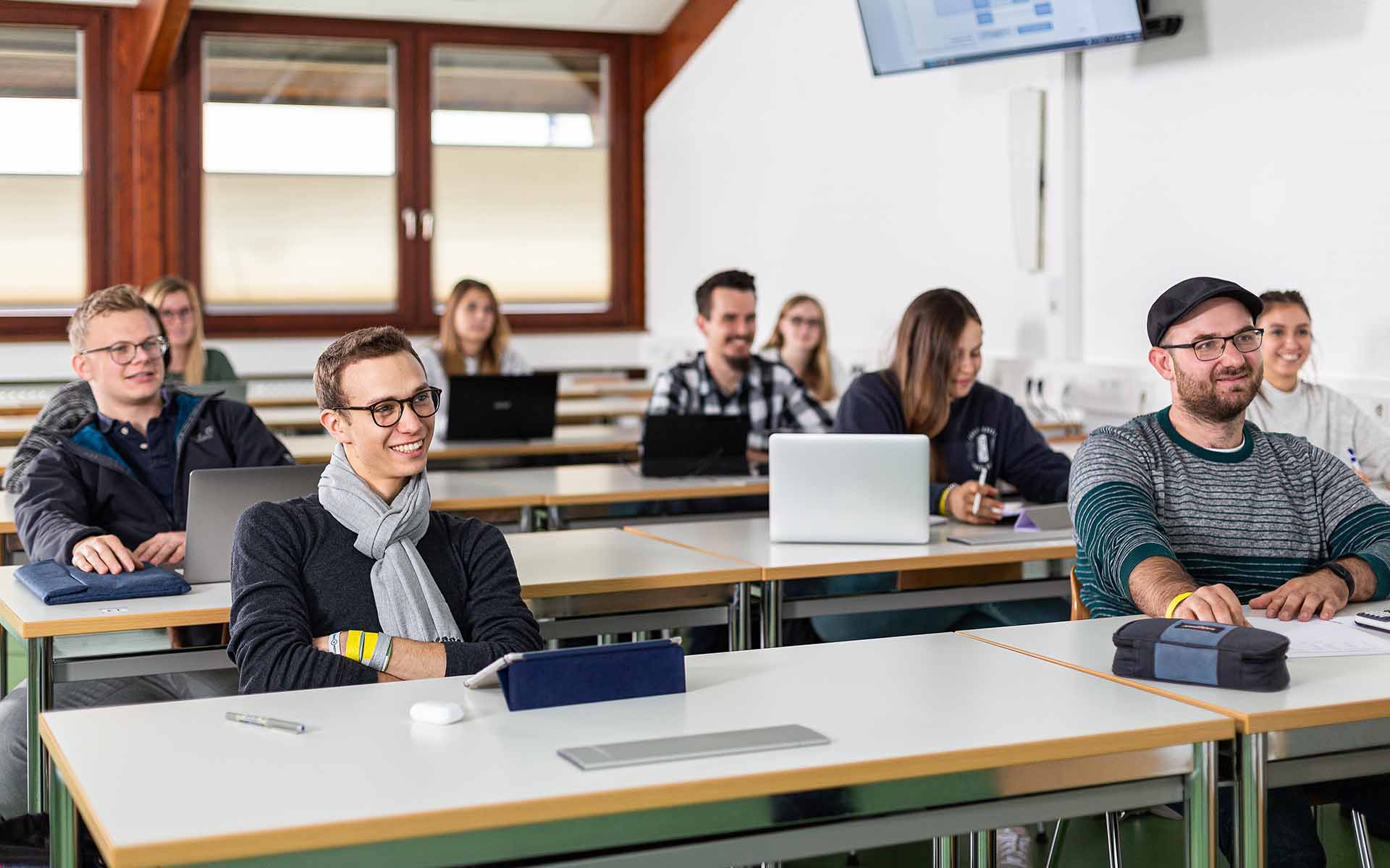 Studierende, die in mehreren Reihen hintereinander an Tischen in einem Hörsaal sitzen und lächelnd nach vorne blicken