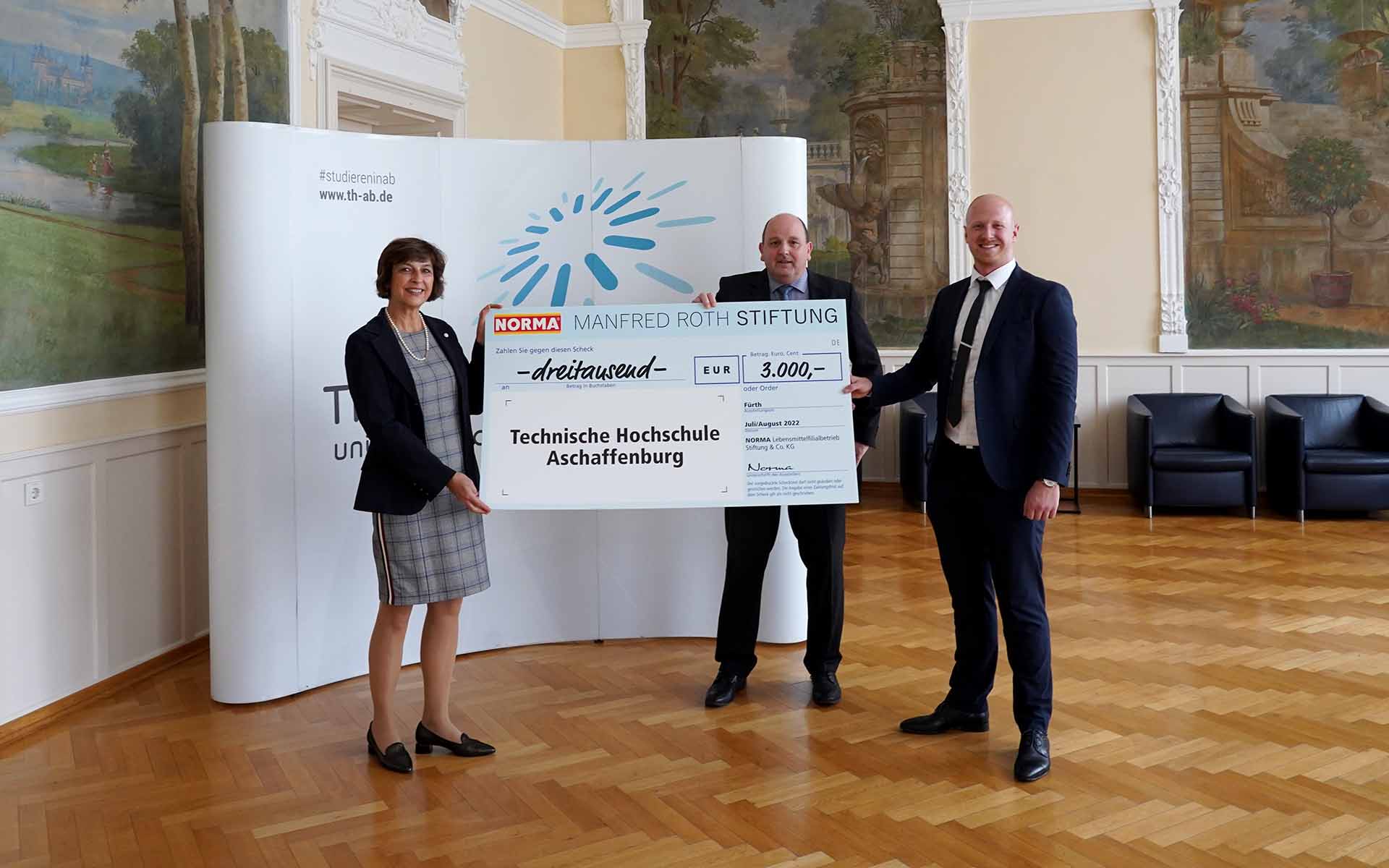 Zwei Männer in schwarzen Anzügen überreichen einer Frau im schicken Kostüm symbolisch einen großformatigen Scheck im Hock-Saal der Hochschule vor einer Wand mit dem TH-Logo