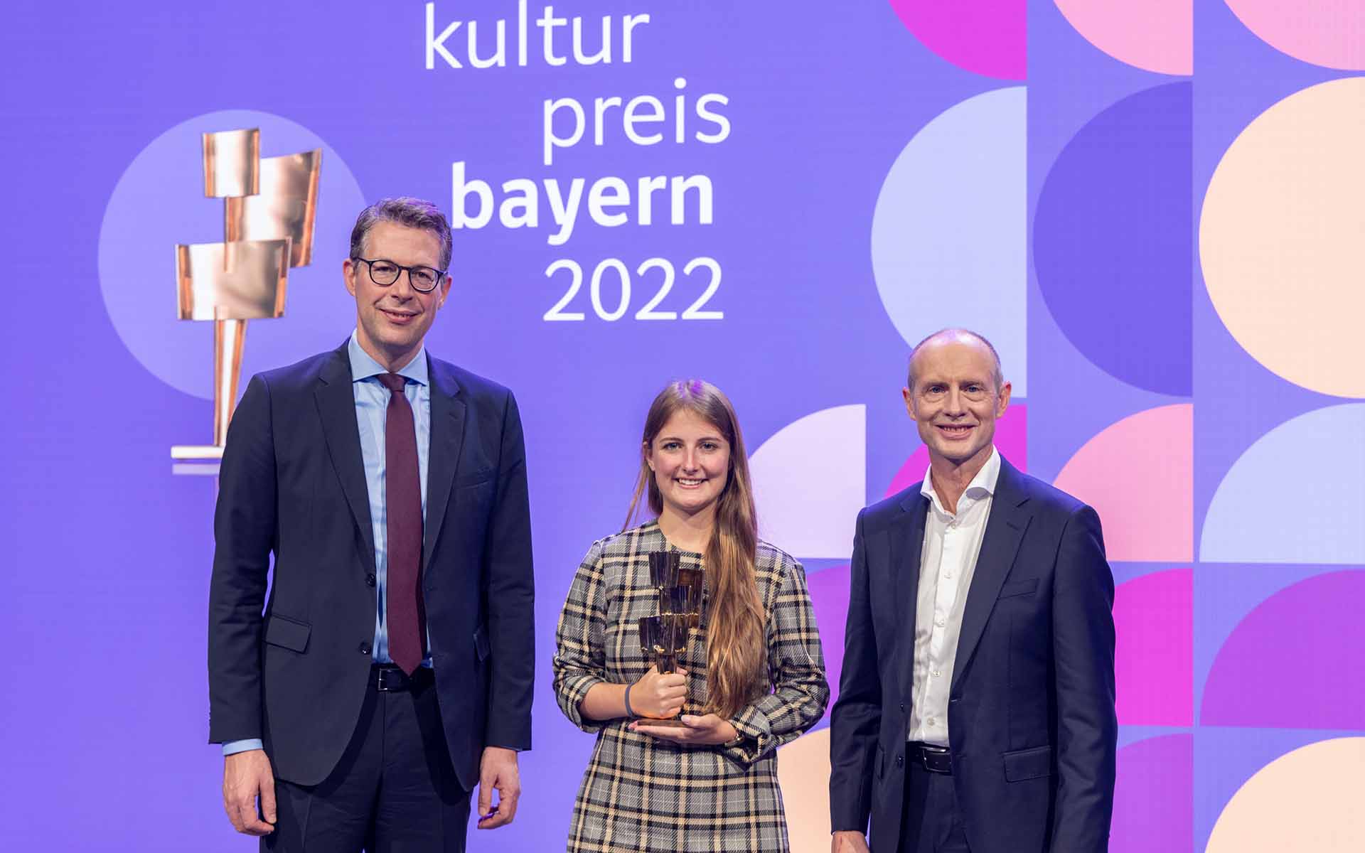 Nebeneinander vor einer violetten Wand mit der Aufschrift Kulturpreis Bayern 2022 steht ein Mann mit dunklem Anzug und Brille, eine junge Frau mit langen dunkelblonden Haaren und einer goldenen Statue in der Hand und ein Mann mit Glatze und ebenfalls dunklem Anzug