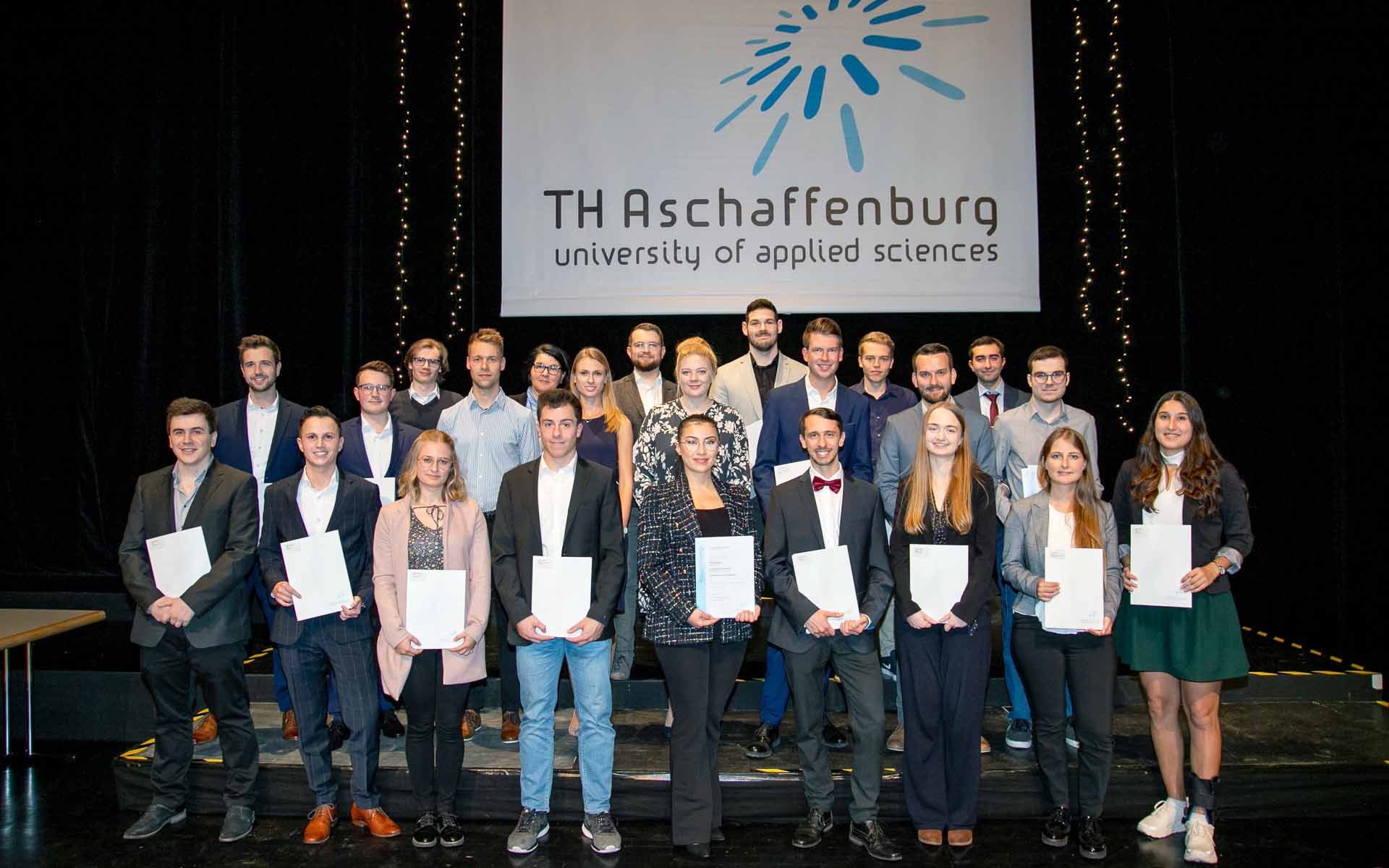 23 Studierende stehen in mehreren Reihen hintereinander mit ihren Zeugnissen in der Hand auf der Bühne vor einem Banner mit dem TH-Logo