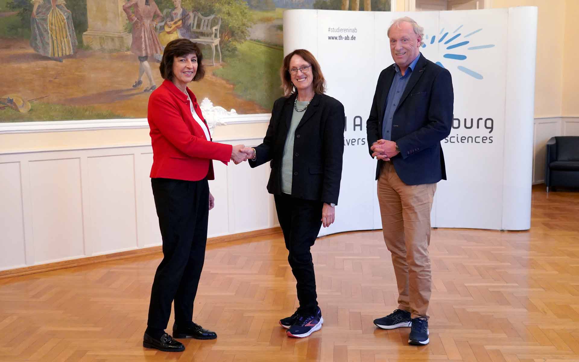 Eine Frau im roten Blazer gibt einer Frau mit Brille und schwarzem Blazer lächelnd die Hand. Rechts daneben steht ein Mann mit blauem Sakko und brauner Hose vor einer Wand mit dem TH-Logo und einem Gemälde im Hocksaal.