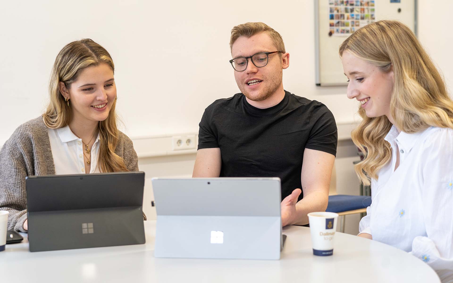 Luisa und zwei ihrer Kommilitonen sitzen vor zwei Laptops und unterhalten sich dabei lachend.