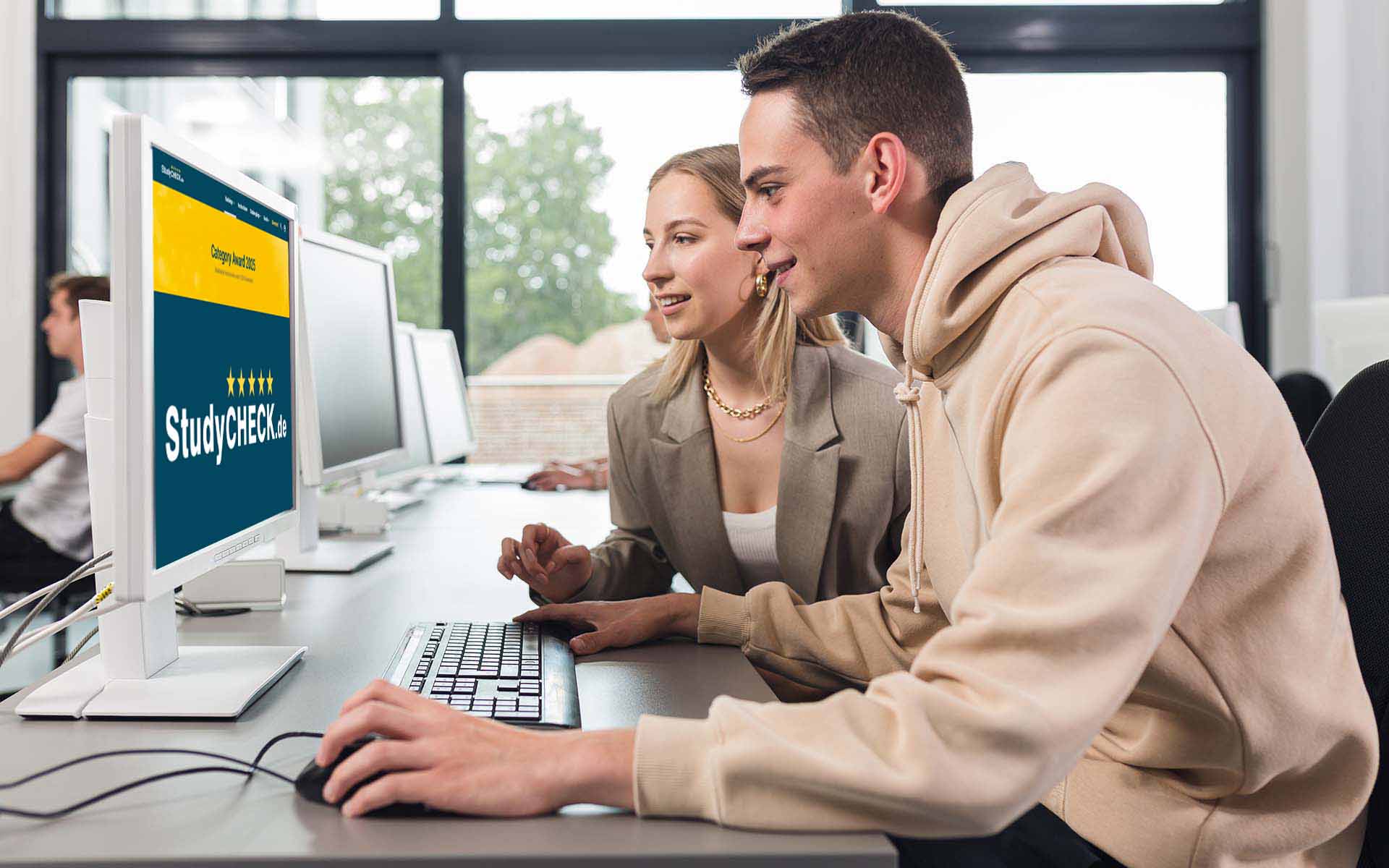 Student und Studentin sitzen vor Bildschirm, auf die die Studycheck-Website mit Logo auf blauem Hintergrund mit gelbem Banner zu sehen ist